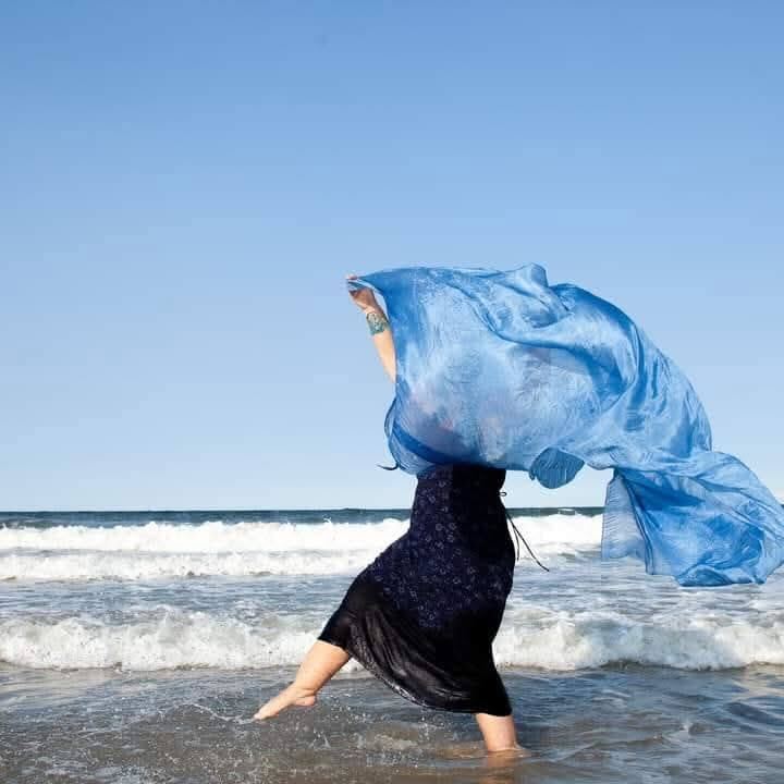 woman dances on a beach holding a blue silk scarf billowing in the wind
