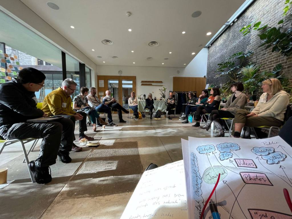 A group sitting on chairs in a circle the workshop room at The Garden Museum, sun streaming in. A hand-drawn diagram pokes up from the bottom right corner